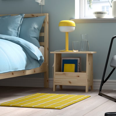 Yellow checked VÄNDPLATS rug beside bed with lamp, compact.