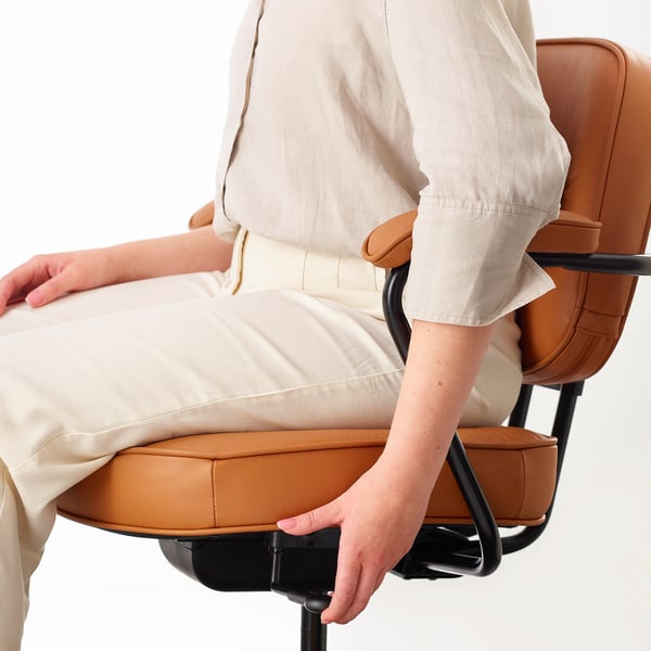 Osoba sedící na ergonomické kancelářské židli s nastavitelnou bederní opěrkou a polstrovanými područkami, čalouněnými kůží.