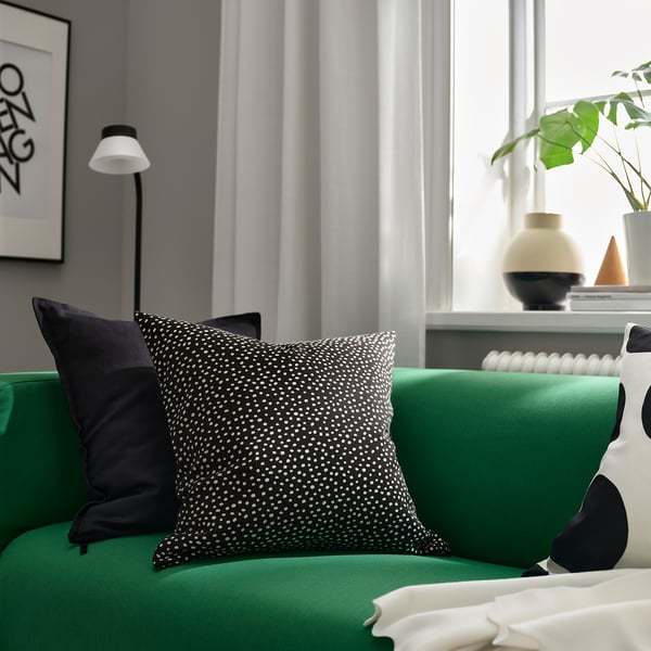 Stylový zelený polštář BJÖRKGRÅMAL s puntíkovaným vzorem na posteli. V kombinaci s černým polštářem, proti šedé stěně s lampou a rostlinou. Moderní výzdoba.