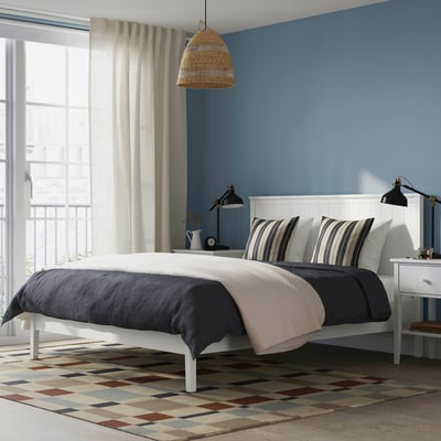 Rám postele GULLABERG. Bílá s dřevěným čelem postele, pruhovanými polštáři, modrá stěna, černá lampa u postele postele.
