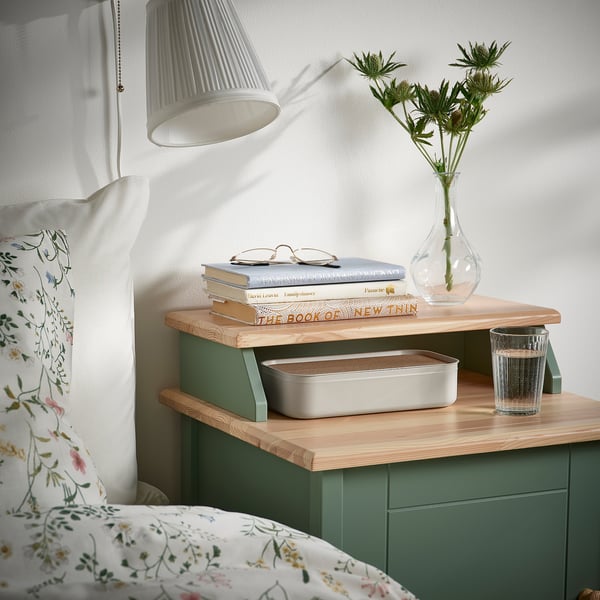 Noční stolek se zeleným organizérem HARVMATTA, který drží knihy, lampu, květiny a sklenici.
