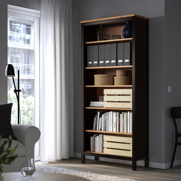 Tmavě hnědá knihovna HEMNES stojí vysoko v rohu, plná knih a úložných košů, vedle šedé pohovky a lampy.