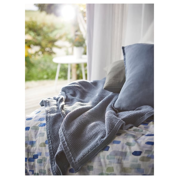 Modrošedá přikrývka na postel s geometrickým potahem. Měkký, recyklovaný polyester. Bílá židle a rostliny viděné oknem, vytvářející útulnou atmosféru.
