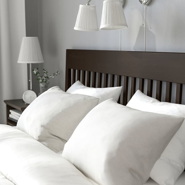 Útulná ložnice s postelí IDANÄS, bílým ložním prádlem, nástěnnými lampami, nočním stolkem a květinami.