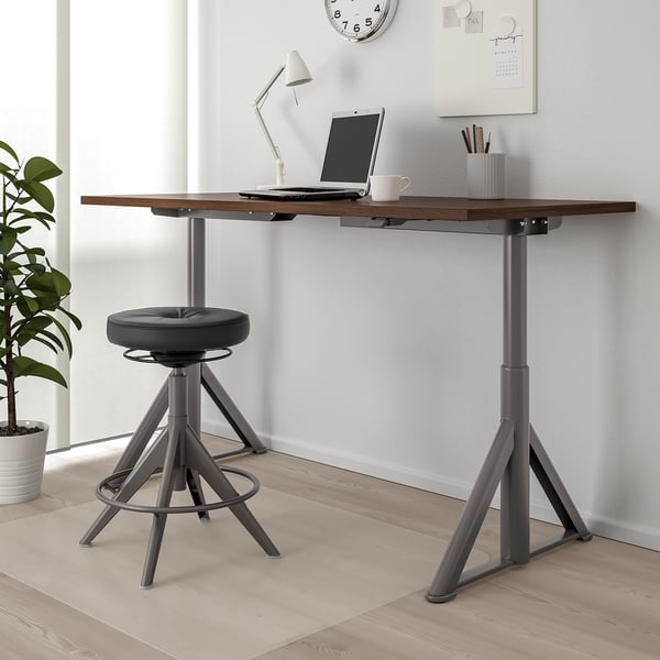 Psací stůl IDÅSEN se sedícím a stojacím stolem s notebookem, lampou a hrnkem. Černá židle pod ní, světlá podlaha, bílé stěny, vedlejší rostlina.