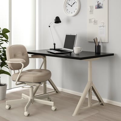 Černý psací stůl IDÅSEN, bílé nohy, béžová židle. Notebook, hrnek, lampa, držák na pero, hodiny, fotografie na zeď.