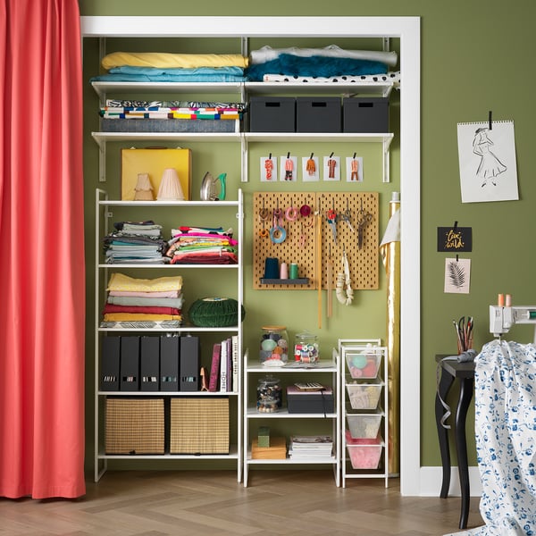 Uspořádaná řemeslná místnost se zelenými stěnami, bílými úložnými policemi JONAXEL, barevnými látkami a uměleckými potřebami. Červený závěs vlevo.