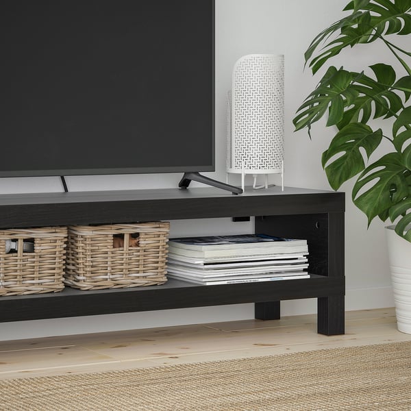ČERNÝ tv stolek s tkanými košíky a naskládanými knihami. Bílá lampa a vedle ní velká zelená rostlina.