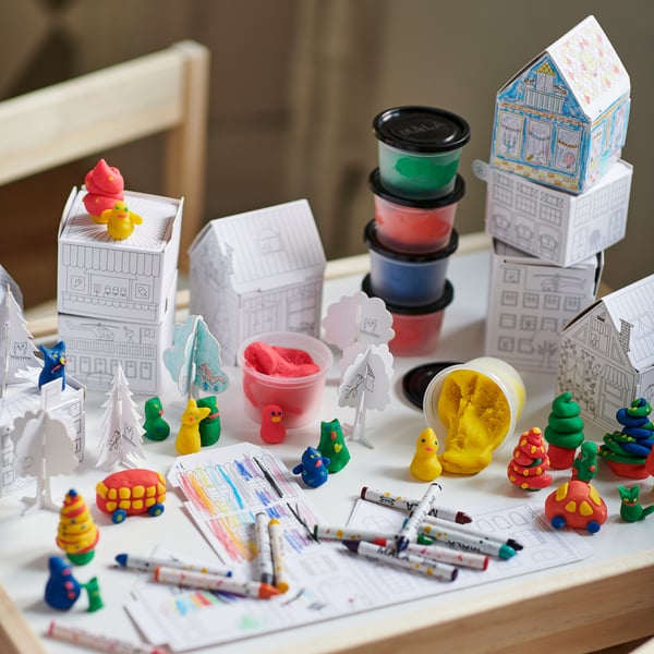 Kreativní herní sestava: barevné papírové domečky, hračky, fixy a modelína na stole, která podporuje nápadité hraní.