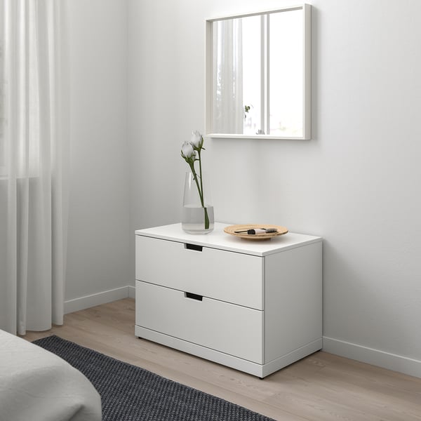 Minimalistický ložnicový kout s komodou, zrcadlem, květinami a podnosem na kosmetiku NORDLI. Měkké světlo přes závěsy.