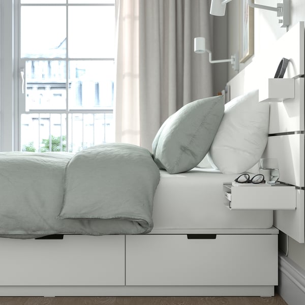 Moderní ložnice s bílým rámem postele NORDLI, modrým ložním prádlem, velkým oknem a úložným prostorem u postele. Zásuvky pod postelí pro uložení.