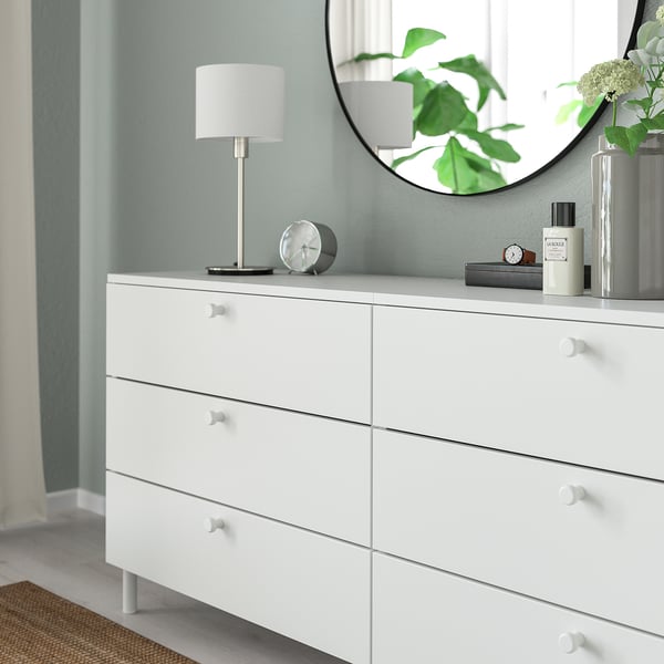 Bílá komoda se šesti zásuvkami, z nichž každá má kulaté knoflíky pro snadné uchopení. Nahoře s kulatým zrcadlem, lampou a dekorativními předměty.