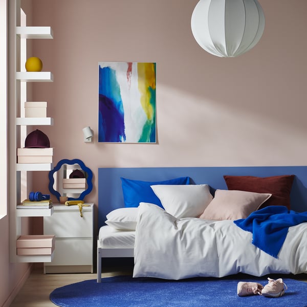 Moderní ložnice s velkou modrou přikrývkou PILTANDVINGE na manželské posteli, odvážným uměním, bílými policemi s barevným dekorem.