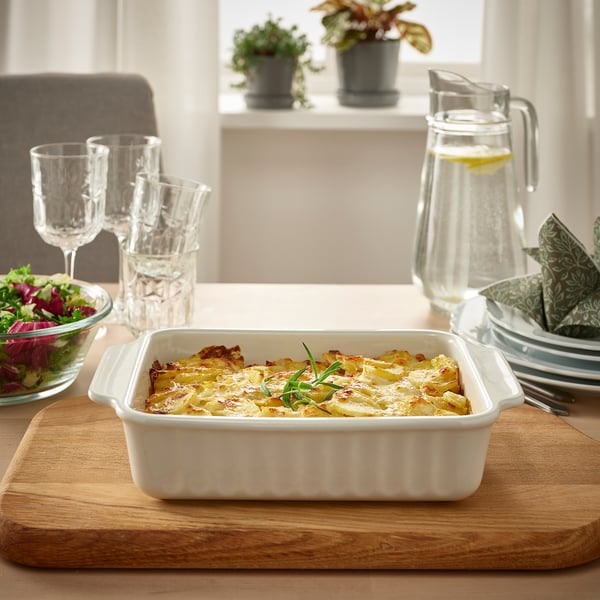 Gratinovaná bílá kamenina na dřevěném prkénku, se sklenicemi, džbánem a salátovou mísou na světlém stole.
