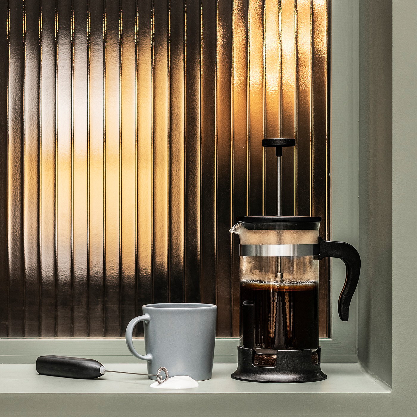 Černý french press kávovar na okenním parapetu s šedým hrnkem, napěňovací hůlkou a sametovou taškou u okna s vertikálními žaluziemi.