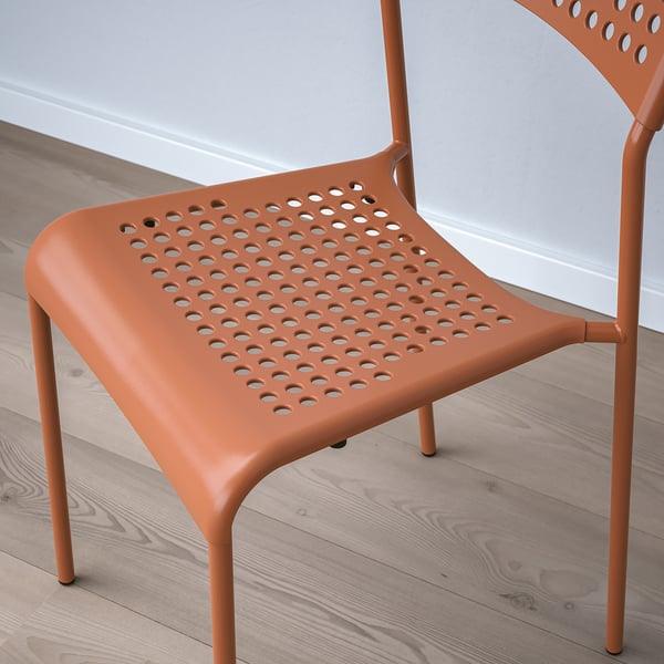 Oranžová moderní židle s perforovaným sedákem a opěradlem, navržená pro stohovatelnost a snadné čištění, umístěná na dřevěné podlaze.