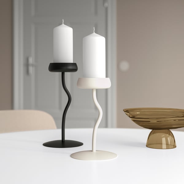 Dva svícny SILVERPÄRON, černý a bílý s bílými svíčkami, elegantně zakřivené, proti béžové židli a mosazné míse na bílém stole.