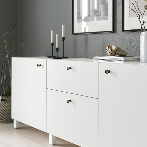 Bílá skříňka s černými úchytkami, s antracitovou povrchovou úpravou a zaoblenými detaily. V horní části jsou umístěny svícny a dekorační předměty.