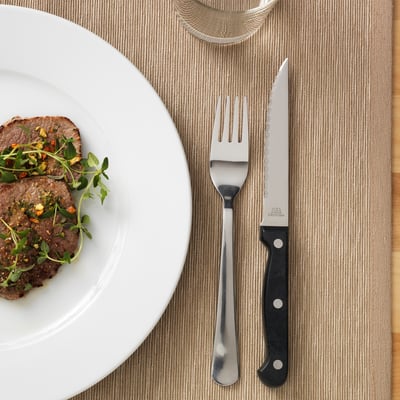 Steakový nůž se zoubkovanou čepelí a černou rukojetí je umístěn vedle vidličky a talíře s vařeným masem ozdobeným zeleninou.