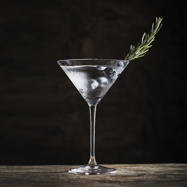 Sklenice na martini se stopkou, obsahující čirou tekutinu a kostky ledu, ozdobená snítkou rozmarýnu.