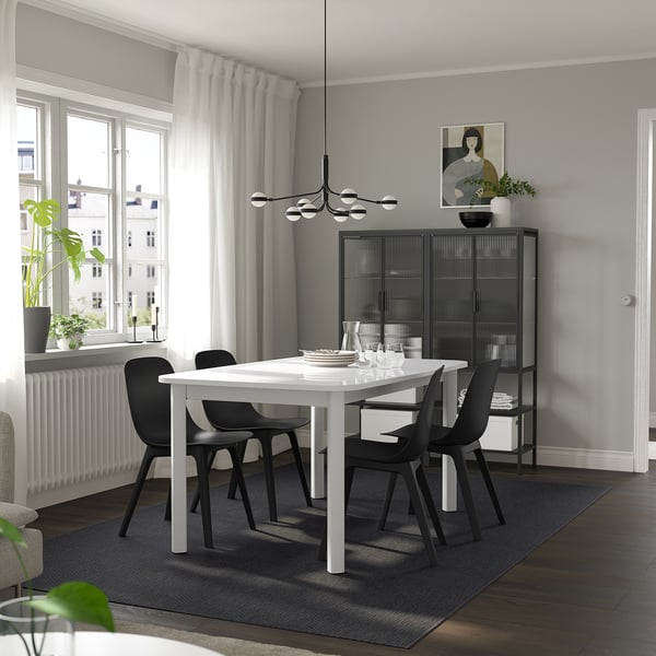 Moderní jídelní kout s bílým stolem STRANDTORP a černými židlemi. Elegantní design kontrastuje s šedými stěnami a velkými okny.