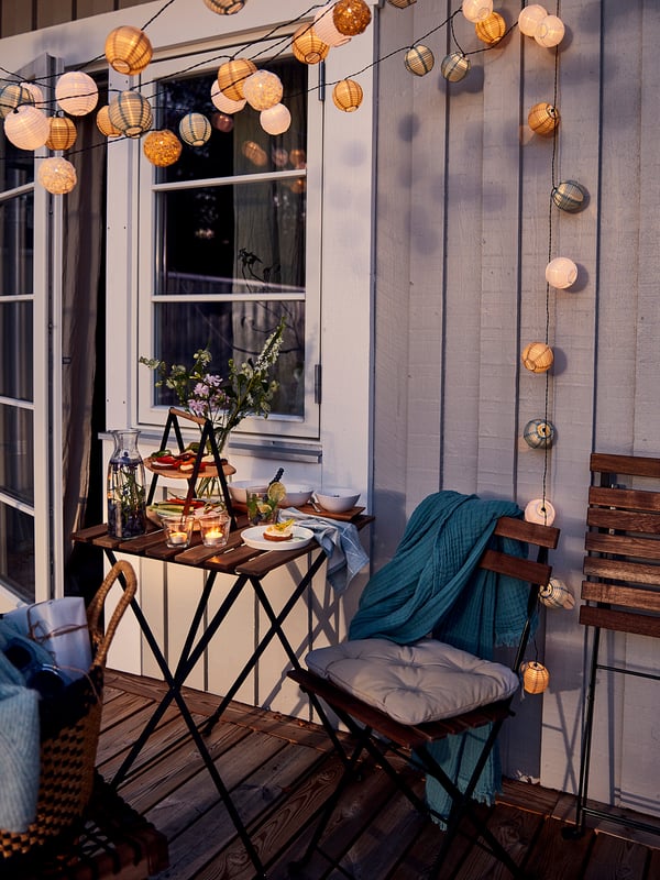 Malý balkon se stolem a židlí TÄRNÖ v černé a dřevěné úpravě, zdobený světelnými řetězy, jídlem a dekou, vytvářející útulné prostředí pro večeři.
