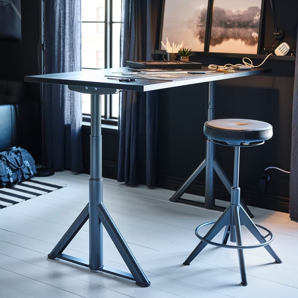 Moderní šedý sedací/stojící stůl s nastavitelnou černou židlí, s kovovými nohami a minimalistickým designem.