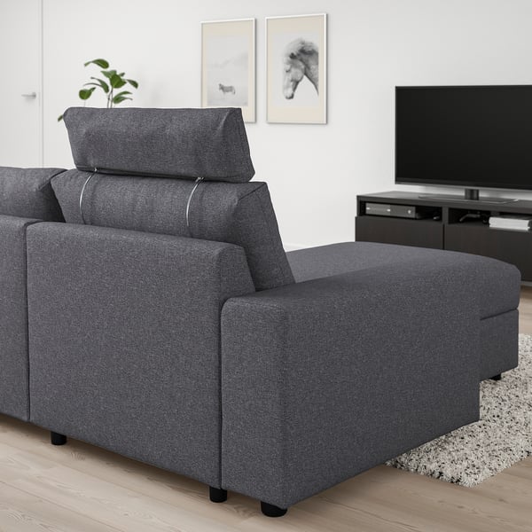 Moderní obývací pokoj s šedou sedací soupravou VIMLE, polohovatelnou opěrkou hlavy, kovovými podpěrami a polyesterovým potahem. Vedle televize, umění a rostliny.
