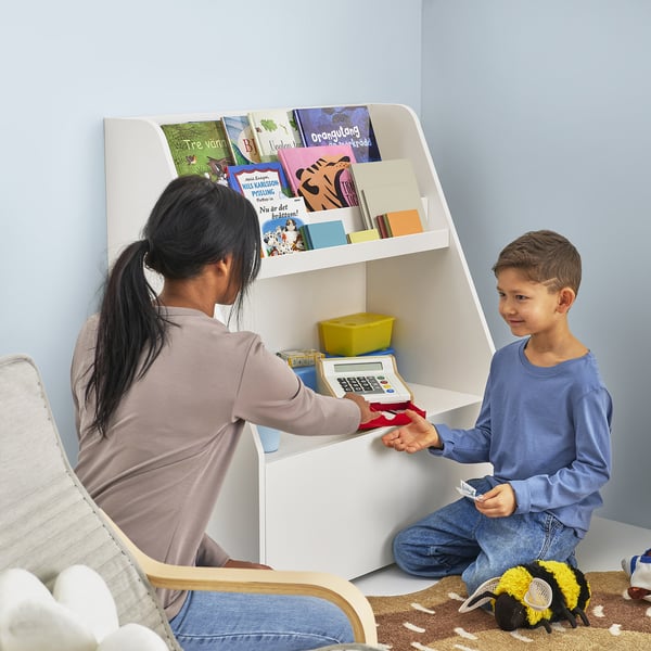 Person assists boy using toy cash register at a child-sized bookcase with shelves and a drawer.