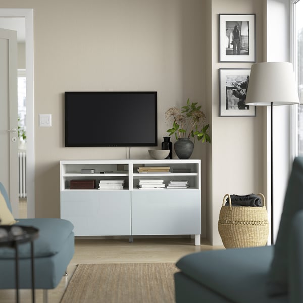 Modern living room with BESTÅ tv unit, blue armchairs, flat-screen tv, books, and decor. Beige wall with pictures, floor lamp, and woven basket.