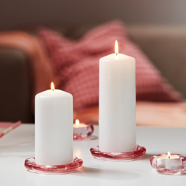 Three lit white candles sit on red glass dishes, creating a flowerbed effect. The dishes and candles are elegantly arranged on a white surface.