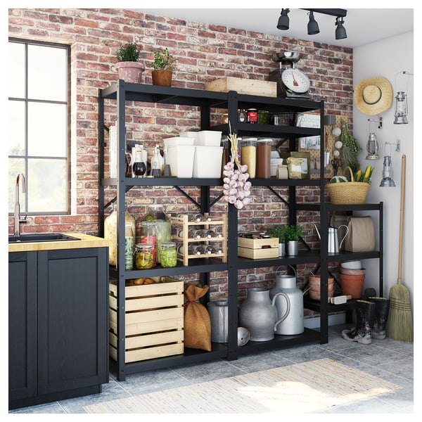 Black metal storage shelves against a brick wall, holding various kitchen items like jars, pots, and utensils, with rustic decor.
