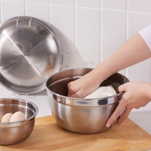 Person mixing dough in a large stainless steel bowl, which nests with similar bowls for space-saving. Metal lids nearby for storing food.