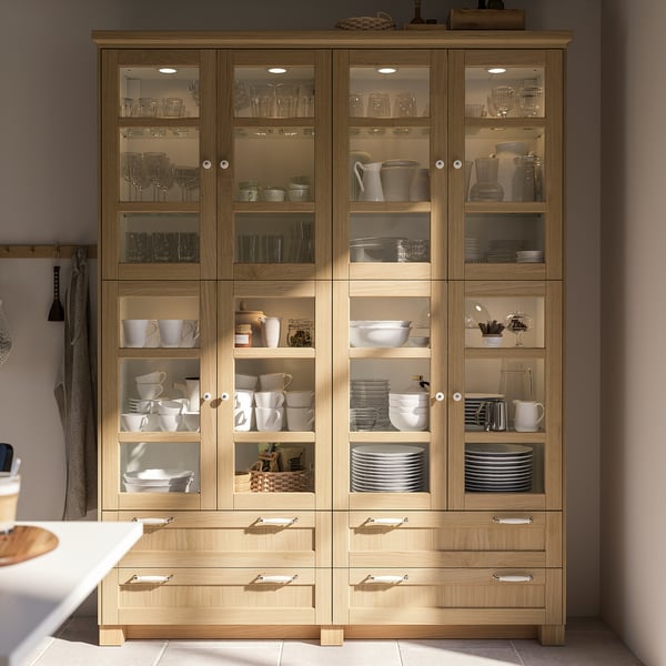 Wooden kitchen cabinet, FORSBACKA design, light brown colour, glass doors, multiple shelves, neatly organised dishes, drawer at the bottom.