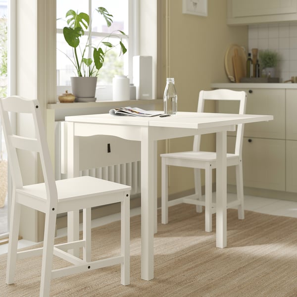 Simple white drop-leaf dining table with two chairs, set near a window with plants.