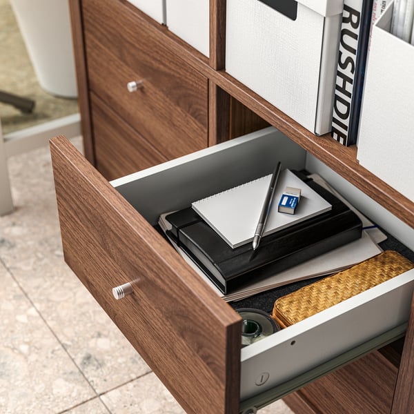 Wooden drawer open with notebook, pens, and other office supplies inside. Part of KALLAX storage system.