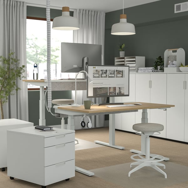 A white sit/stand desk in a modern home office with wooden top, cable management, and ergonomic chair.