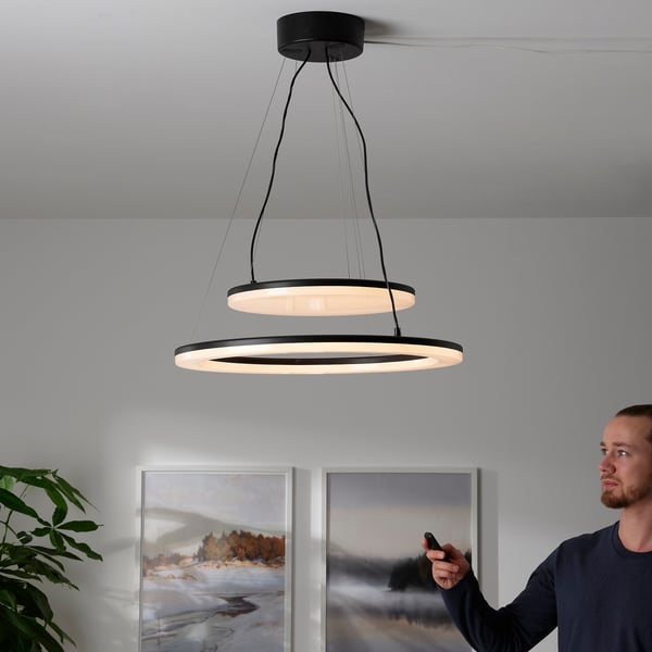 Person adjusting modern dual-ring pendant light with remote, controlling brightness and positioning.