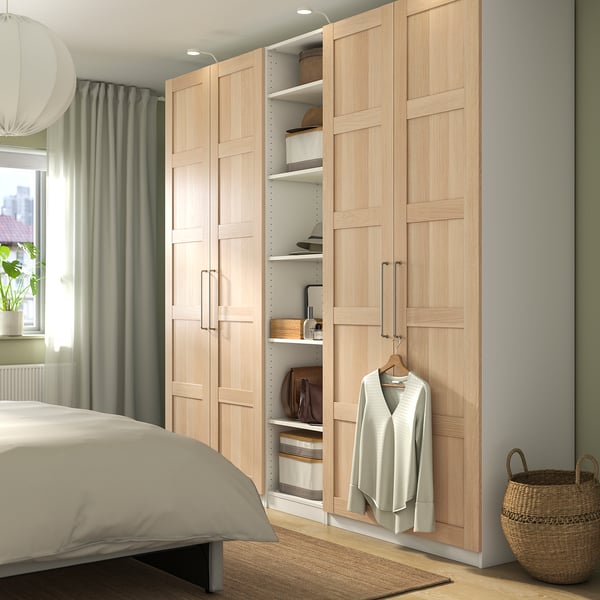 Wooden wardrobe beside bed, one door open showing clothes and shelves. Soft grey room with large window and plant.