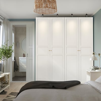 Modern bedroom with large white PAX wardrobe. Gold handles, dark bedding, wicker light, bedside lamp, and plant by door.
