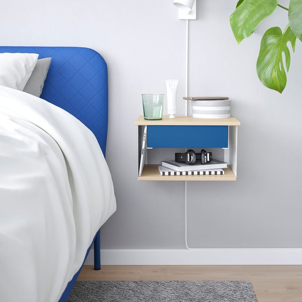 Wall-mounted STOMSÖ bedside table in blue and wood with open/closed storage, holding glass, books, lamp, and plant.