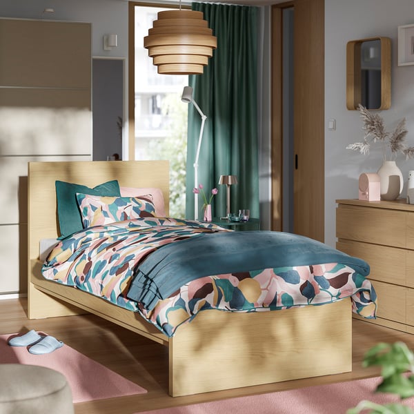 Low light wood bed with colourful bedding, dark green curtains, pink rug, and wooden furniture.