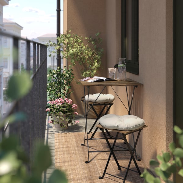 Small wooden & black metal stools with cushions on a balcony, next to potted plants and a table with a book and drinks.