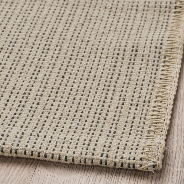 Flatwoven cotton rug, beige with small dark dots, neatly bordered, laid on wooden floor.