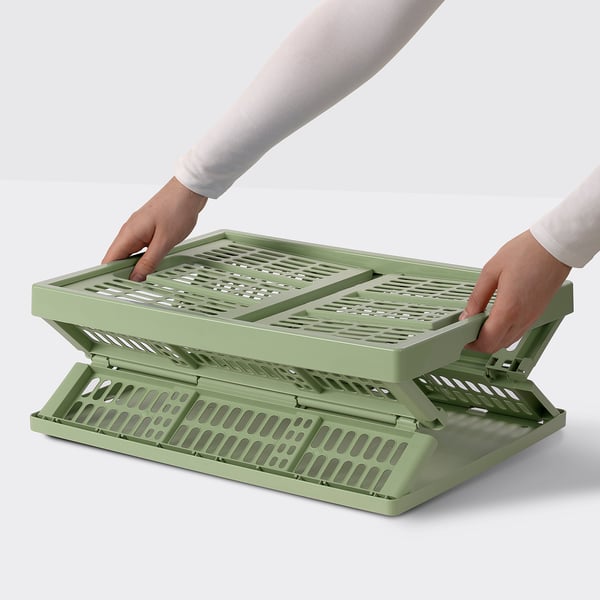 Person holding an olive green VEDKLYV folding box with handles, demonstrating how to unfold it effortlessly.