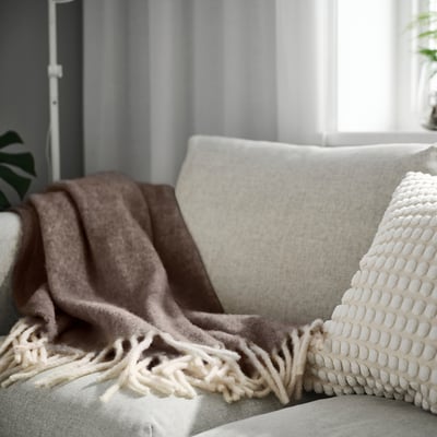 ALLFRAKEN graue Wolldecke mit Fransen auf beigem Sofa, schmückt großen Raum.