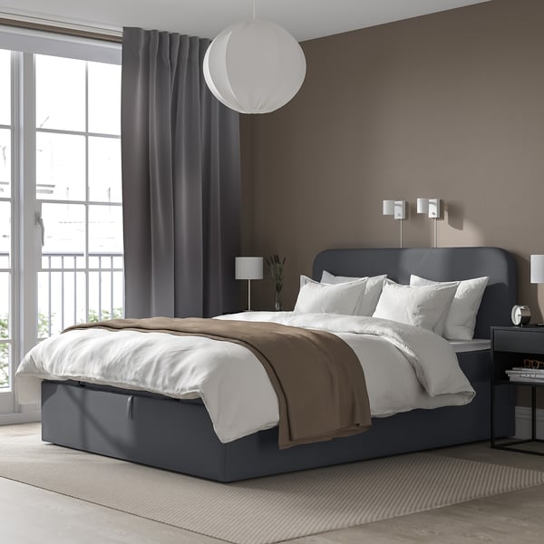 Modernes graues Schlafsofa mit Stauraum, weißen Kissen, grauen Vorhängen, Hängelampe, Beistelltischen mit Lampen und einem Teppich vor einer braunen Wand.