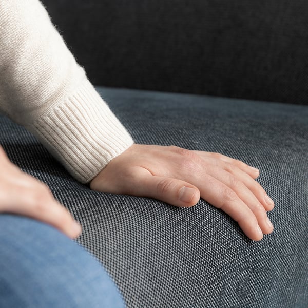 Die Hand ruht auf einem grauen Stoffsofa. Die Person trägt einen weißen Pullover und blaue Jeans.