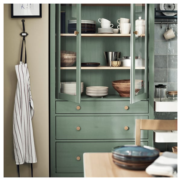 Grüner HEMNES Holzschrank mit Glastüren, in dem Geschirr und Schalen ausgestellt sind. Die Schürze hängt auf der linken Seite, die Küchentheke im Vordergrund.