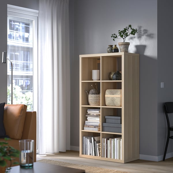KALLAX Holzregal mit Büchern, Pflanzen, Dekor. Brauner Ledersessel, Fenster mit Vorhängen.